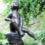 Niño en estatua de bronce junto a una fuente, rodeado de naturaleza y simbolizando energía vital