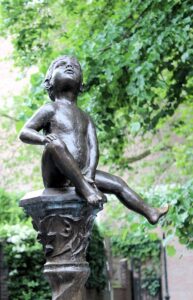 Niño en estatua de bronce junto a una fuente, rodeado de naturaleza y simbolizando energía vital